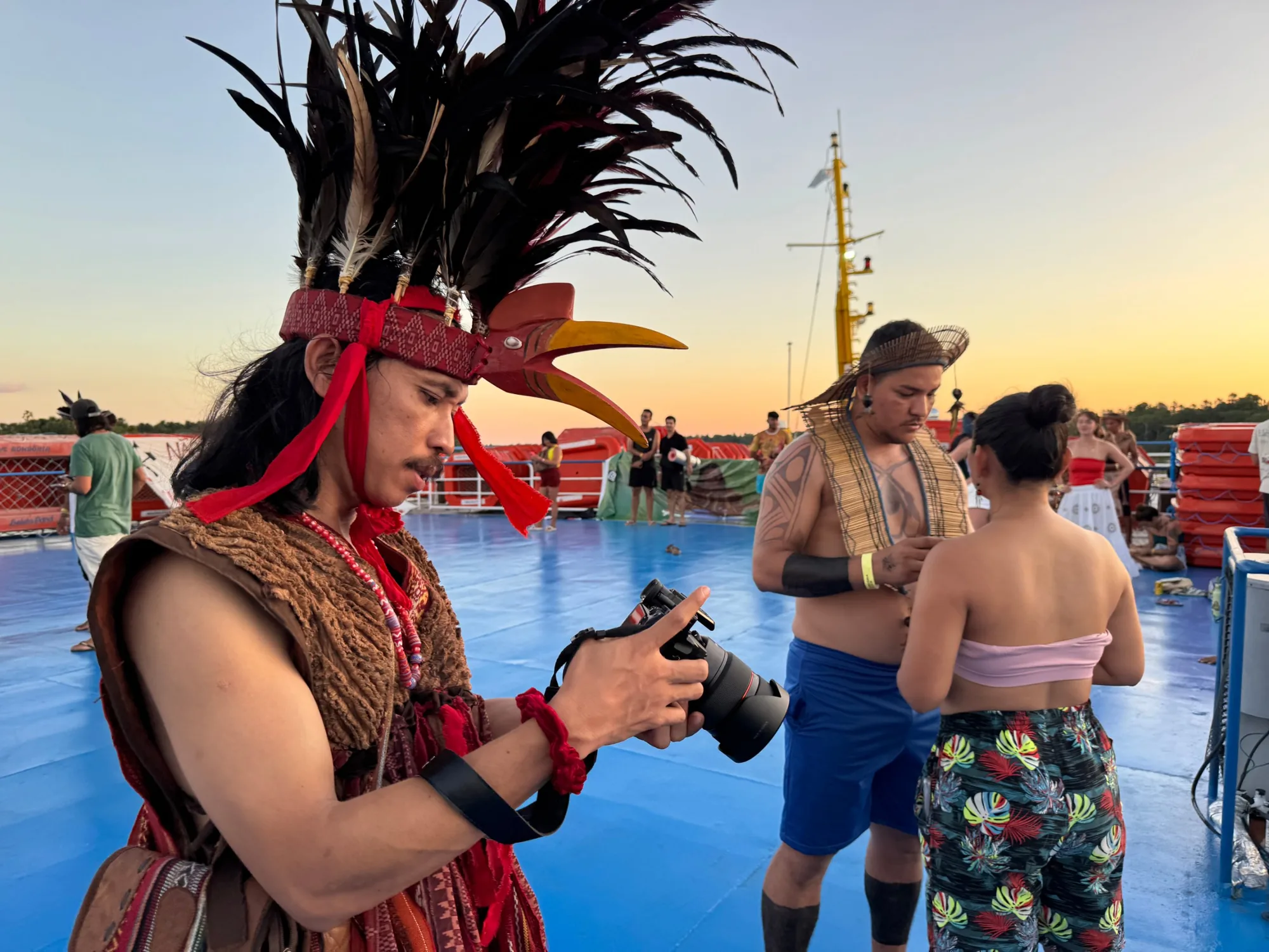 Un realizador con tocado de plumas revisa su cámara en la cubierta del barco al atardecer